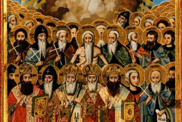 Ένα Πυκνό, Σχεδόν Ασφυκτικά Γεμάτο Πλήθος Φωτοστεφάνων Καταλαμβάνει Το Μεγαλύτερο Μέρος Αυτής Της Φορητής Εικόνας, Γραμμένης Το 1810 Στη Μονή Βατοπεδίου Του Αγίου Όρους. Δεν Πρόκειται Για Την Απεικόνιση Ενός Μεμονωμένου Προσώπου Ούτε Για Μια Συνηθισμένη Δεησιακή Σύνθεση, Αλλά Για Κάτι Πιο Φιλόδοξο: Μια Συλλογική Εικόνα Των Αγίων Πάντων Αγίου Όρους, Όλων Δηλαδή Των Μοναχών, Ασκητών Και Ιεραρχών Που Έζησαν Και Αγίασαν Στη Χερσόνησο Του Άθω, Γνωστών Με Το Όνομά Τους Αλλά Και Άγνωστων, Όσων Η Μνήμη Διασώθηκε Μόνο Ως Συλλογικό Σώμα. Ο Ζωγράφος Παραμένει Ανώνυμος, Όπως Συμβαίνει Με Το Μεγαλύτερο Μέρος Της Αγιορειτικής Εργαστηριακής Παραγωγής Της Περιόδου, Όμως Η Χρονολόγηση Στο 1810 Είναι Σαφής Και Συνδέει Το Έργο Με Μια Συγκεκριμένη Στιγμή Της Αθωνικής Πνευματικότητας, Λίγα Μόλις Χρόνια Μετά Τη Σύνθεση Του Εγκωμιαστικού Λόγου Του Νικοδήμου Αγιορείτη Για Τους Πατέρες Του Άθω. Στο Πάνω Τμήμα Δεσπόζει Η Ένθρονη Θεοτόκος Με Τον Χριστό, Ενώ Από Κάτω Απλώνεται, Σε Διαδοχικές Σειρές, Το Αμέτρητο Πλήθος Των Οσίων Μορφών. Η Δουλειά Αυτή Εντάσσεται Στη Διεργασία Μέσω Της Οποίας Το Άγιον Όρος Οργάνωσε Την Ίδια Του Τη Μνήμη Σε Εικαστική Μορφή, Κάτι Που Η Μονή Βατοπεδίου Εξακολουθεί Να Τεκμηριώνει Και Σήμερα Μέσα Από Το Αρχείο Και Τη Βιβλιοθήκη Της. Η Ανάλυση Που Ακολουθεί Εξετάζει Τη Σύνθεση Αυτή Ως Ιστορικό Τεκμήριο Μιας Ζωγραφικής Παράδοσης, Με Έμφαση Στη Δομή, Στα Χρώματα Και Στις Επιγραφές Της.