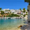 Μικρό λιμάνι στην Αλόννησο με δεμένες βάρκες, λευκά σπίτια στην πλαγιά και ψηλό στρωματωμένο βράχο στα δεξιά.