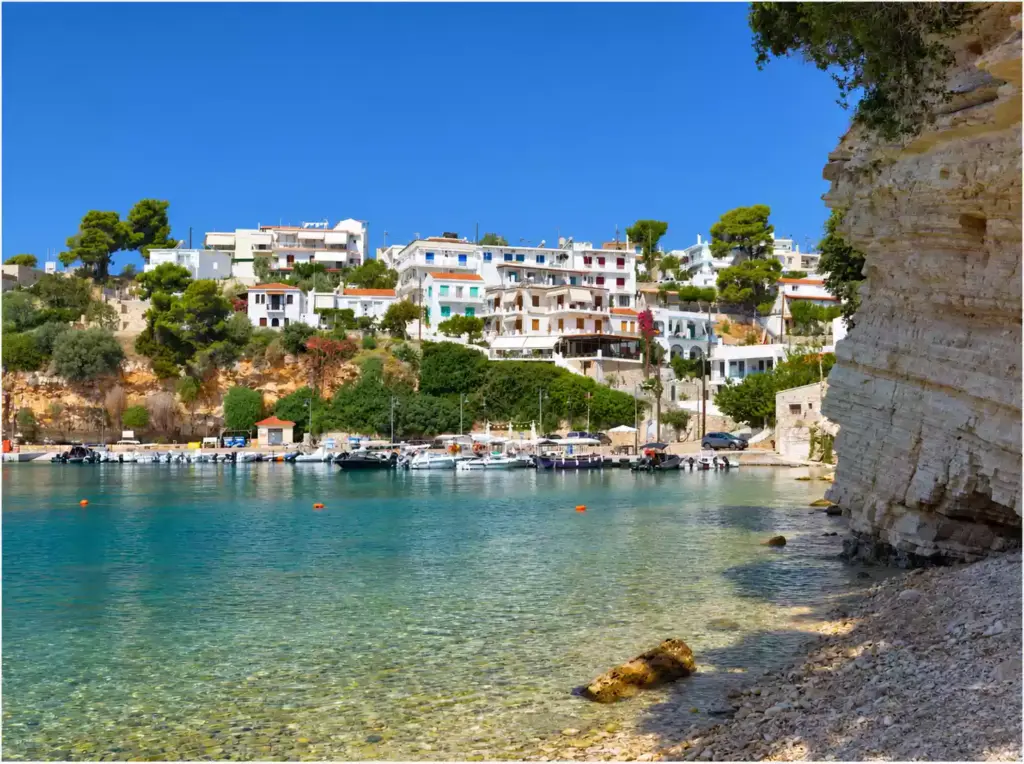Μικρό λιμάνι στην Αλόννησο με δεμένες βάρκες, λευκά σπίτια στην πλαγιά και ψηλό στρωματωμένο βράχο στα δεξιά.