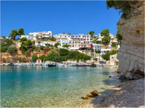 Μικρό λιμάνι στην Αλόννησο με δεμένες βάρκες, λευκά σπίτια στην πλαγιά και ψηλό στρωματωμένο βράχο στα δεξιά.