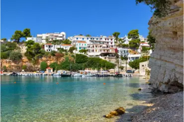 Μικρό λιμάνι στην Αλόννησο με δεμένες βάρκες, λευκά σπίτια στην πλαγιά και ψηλό στρωματωμένο βράχο στα δεξιά.