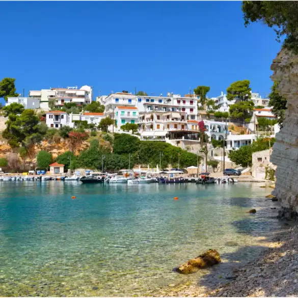 Μικρό λιμάνι στην Αλόννησο με δεμένες βάρκες, λευκά σπίτια στην πλαγιά και ψηλό στρωματωμένο βράχο στα δεξιά.