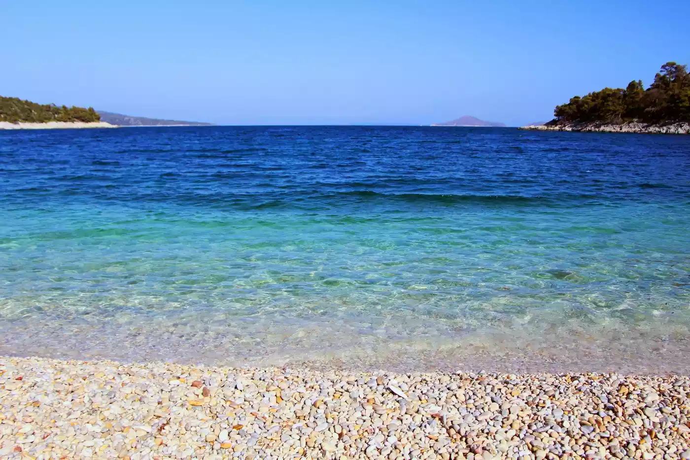 Ανοιχτόχρωμα βότσαλα και διαφανή γαλαζοπράσινα νερά στην Αλόννησο, με πευκόφυτες βραχώδεις ακτές στις άκρες του κόλπου.
