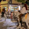 Γυναίκα με παιδί και άνδρας πάνω σε στολισμένα λευκά άλογα, δίπλα σε τραπέζια καταστημάτων σε νυχτερινό πλακόστρωτο.