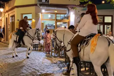 Γυναίκα με παιδί και άνδρας πάνω σε στολισμένα λευκά άλογα, δίπλα σε τραπέζια καταστημάτων σε νυχτερινό πλακόστρωτο.