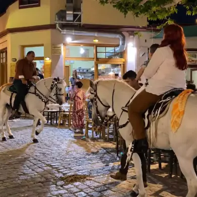 Γυναίκα με παιδί και άνδρας πάνω σε στολισμένα λευκά άλογα, δίπλα σε τραπέζια καταστημάτων σε νυχτερινό πλακόστρωτο.