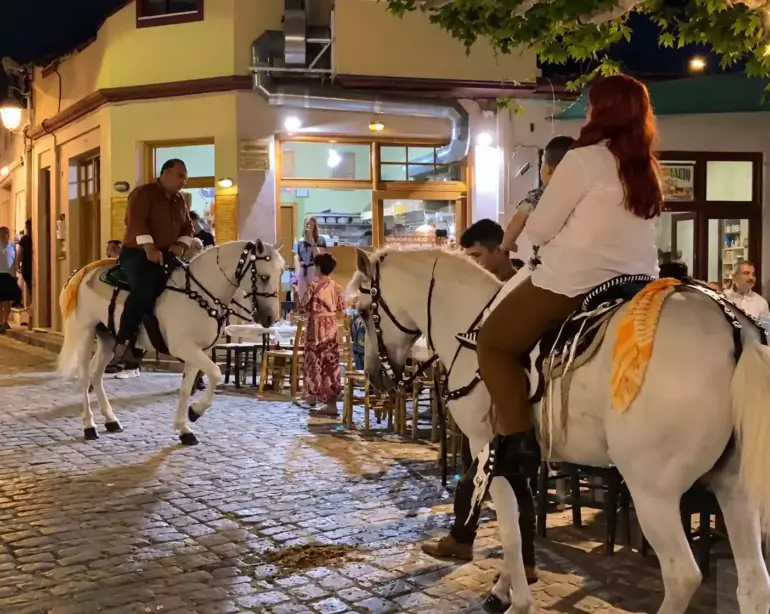 Γυναίκα με παιδί και άνδρας πάνω σε στολισμένα λευκά άλογα, δίπλα σε τραπέζια καταστημάτων σε νυχτερινό πλακόστρωτο.