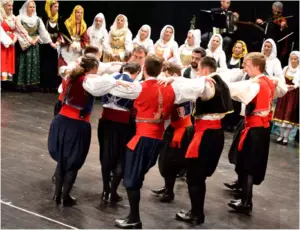 Άνδρες με βράκες και κόκκινα ζωνάρια χορεύουν πιασμένοι από τους ώμους, με γυναίκες και μουσικούς στο βάθος.