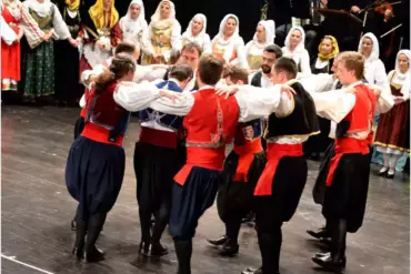 Männer in Pluderhosen und roten Schärpen tanzen, die Hände auf den Schultern der Nachbarn, dahinter Frauen und Musiker.