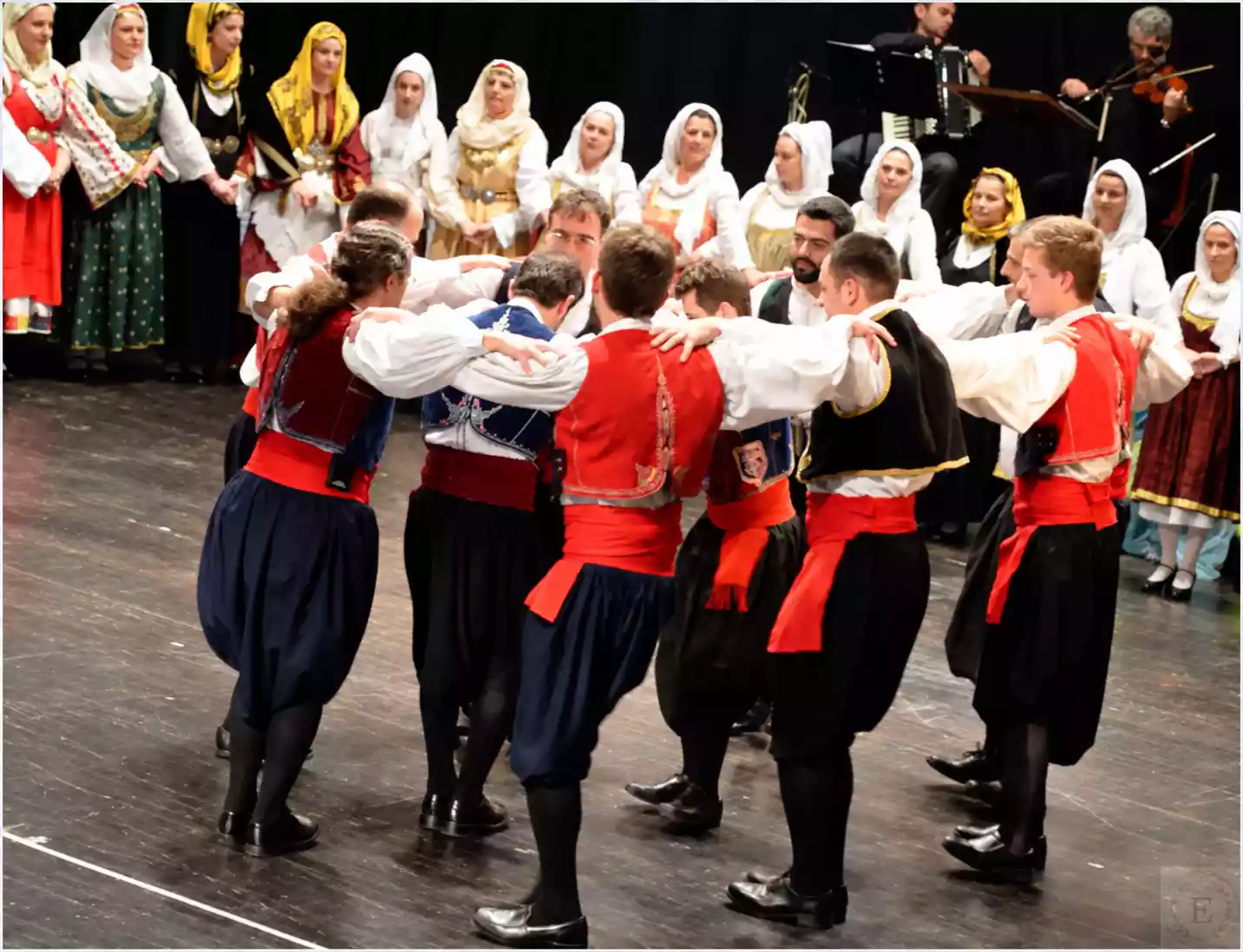 Männer in Pluderhosen und roten Schärpen tanzen, die Hände auf den Schultern der Nachbarn, dahinter Frauen und Musiker.