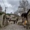Empedrado entre edificios de piedra, con un vano en arco a la izquierda y un cobertizo de madera junto a un gran árbol caducifolio a la derecha.