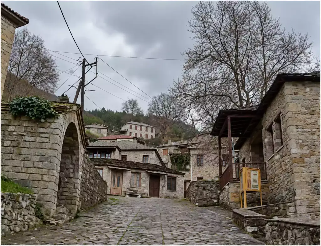Λιθόστρωτο ανάμεσα σε πέτρινα κτίρια, με τοξωτό άνοιγμα αριστερά και ξύλινο στέγαστρο δίπλα σε μεγάλο φυλλοβόλο δέντρο δεξιά.