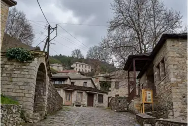 Selciato tra edifici in pietra, con un’apertura ad arco a sinistra e una tettoia in legno accanto a un grande albero a foglia caduca a destra.