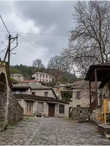 Λιθόστρωτο ανάμεσα σε πέτρινα κτίρια, με τοξωτό άνοιγμα αριστερά και ξύλινο στέγαστρο δίπλα σε μεγάλο φυλλοβόλο δέντρο δεξιά.