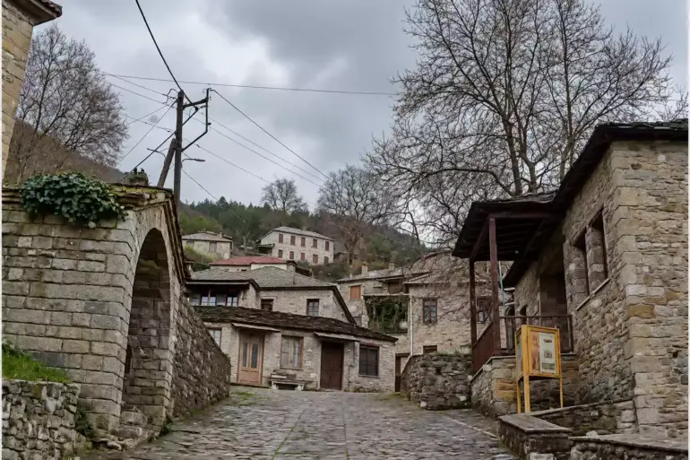 Калдъръмена уличка между каменни сгради, с арковиден отвор вляво и дървен навес до голямо широколистно дърво вдясно.