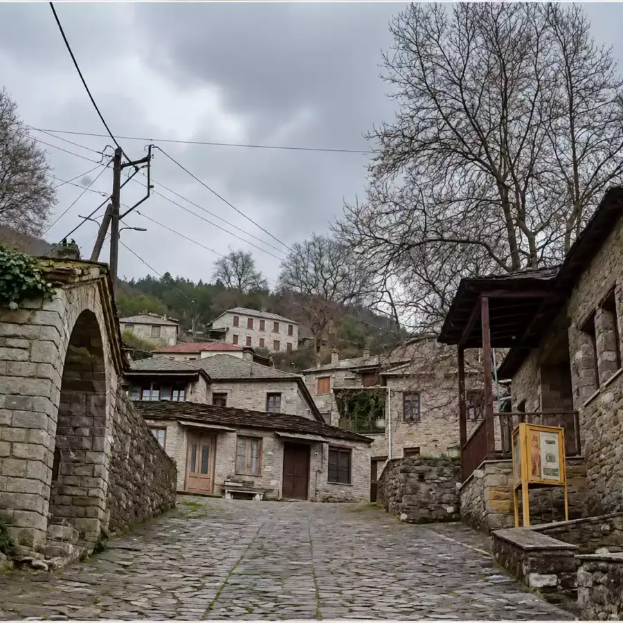 Λιθόστρωτο ανάμεσα σε πέτρινα κτίρια, με τοξωτό άνοιγμα αριστερά και ξύλινο στέγαστρο δίπλα σε μεγάλο φυλλοβόλο δέντρο δεξιά.