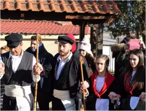 Άνδρες με τραγιάσκες, λευκά ζωνάρια και μαγκούρες δίπλα σε νεαρές χορεύτριες με κόκκινα μαντίλια στους ώμους και λευκές ποδιές.