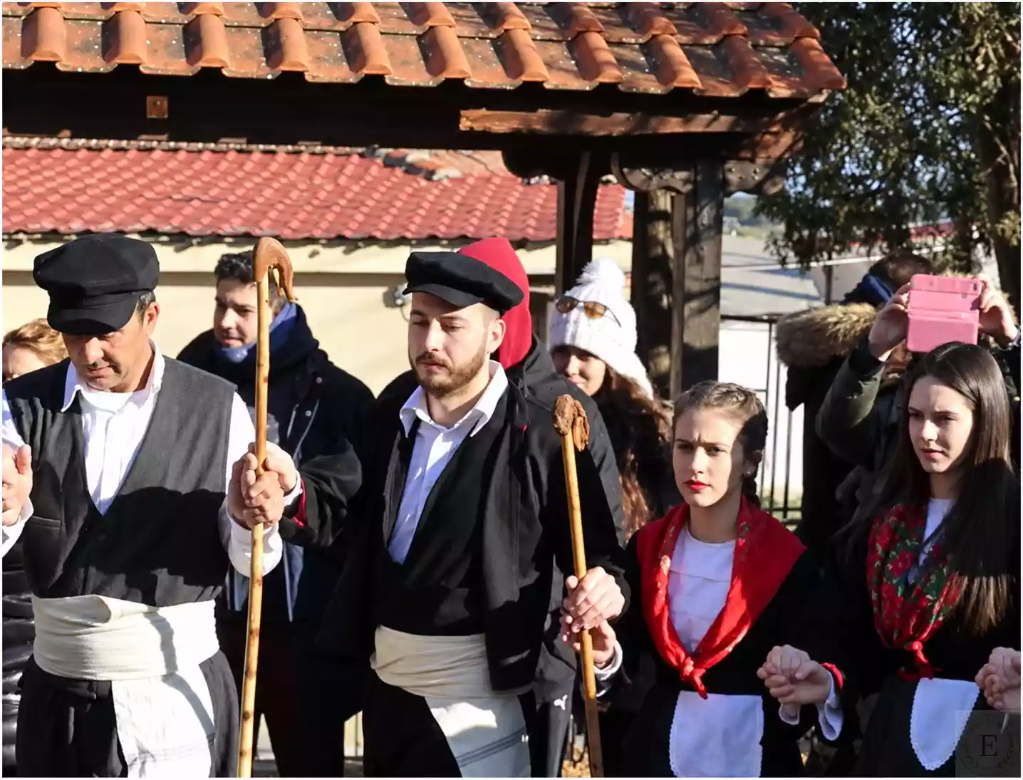 Men in flat caps with white sashes and wooden crooks beside young female dancers wearing red shawls over their shoulders and white aprons.