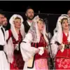 Beyaz, siyah ve kırmızı geleneksel giysiler içinde mikrofonlar önünde şarkı söyleyen kadınlar ve erkekler.