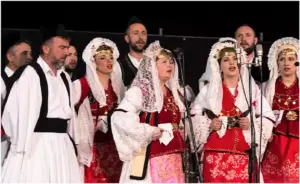 Άνδρες και γυναίκες τραγουδούν μπροστά σε μικρόφωνα, με λευκές, μαύρες και κόκκινες παραδοσιακές ενδυμασίες.