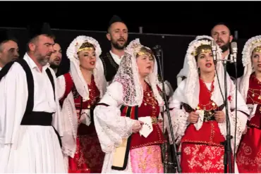 Men and women singing at microphones, dressed in white, black and red traditional costumes.