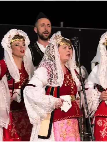 Άνδρες και γυναίκες τραγουδούν μπροστά σε μικρόφωνα, με λευκές, μαύρες και κόκκινες παραδοσιακές ενδυμασίες.