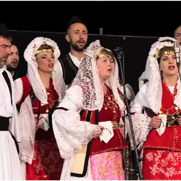 Männer und Frauen singen an Mikrofonen, in weißen, schwarzen und roten Trachten.