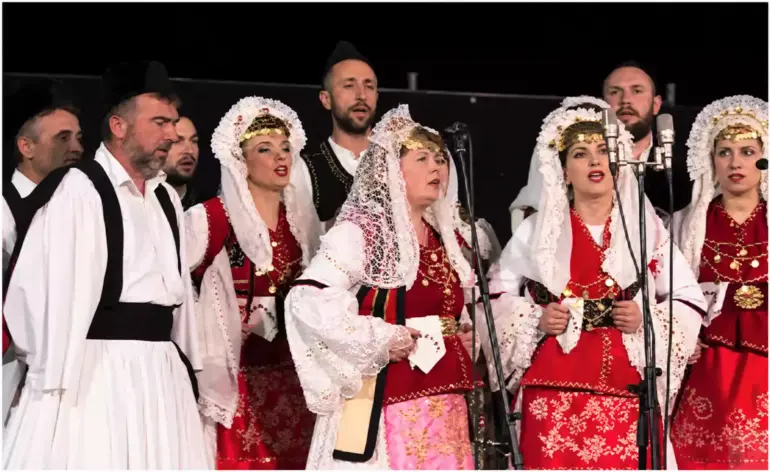 Άνδρες και γυναίκες τραγουδούν μπροστά σε μικρόφωνα, με λευκές, μαύρες και κόκκινες παραδοσιακές ενδυμασίες.