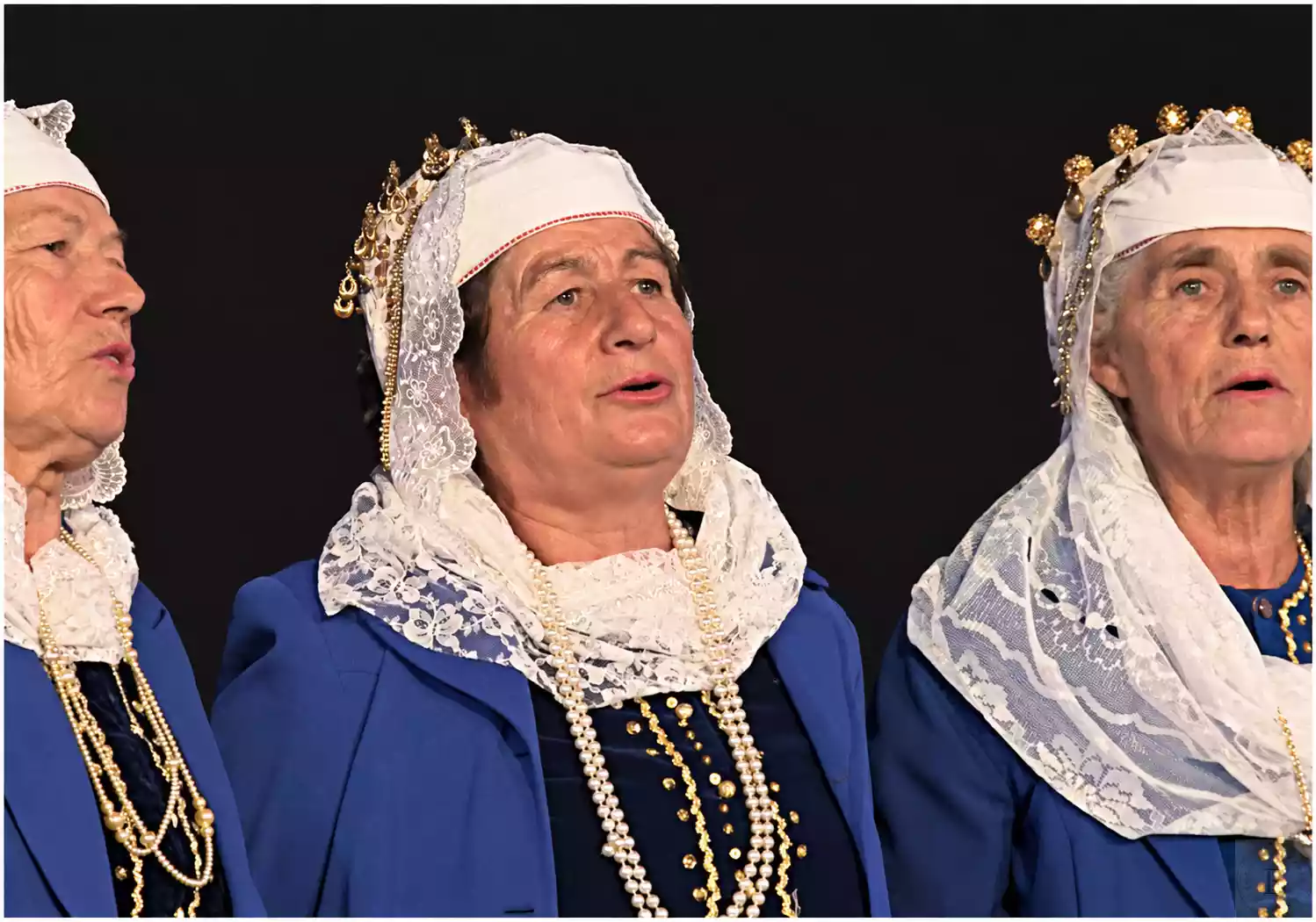 Trois femmes chantant en costumes bleus, coiffes de dentelle blanche et rangs de colliers.
